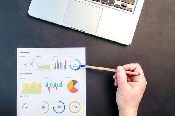 Hand holding a pencil over a printed page of financial charts next to a laptop